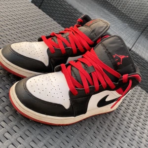 Nike Air Jordan 1 Mid svart/vit/röd - Säljer ett par Nike Air Jordan 1 Mid med klassisk svart, vit och röd färgkombination. Skorna har röda snören, svart Swoosh och detaljer, samt vit tå och sidor. Ovandelen är i skinn och sulan är röd undertill. Perfekta för dig som gillar streetstyle och sneakers med ikonisk design.