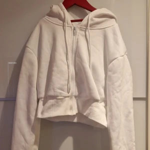 Vit croppad hoodie med texten 'angel' - Säljer en vit croppad hoodie med dragkedja och huva. På ryggen står det 'angel' i stora bokstäver av svarta nitar. Hoodien har långa ärmar och ribbade muddar. Perfekt för dig som vill ha en cool och bekväm stil.