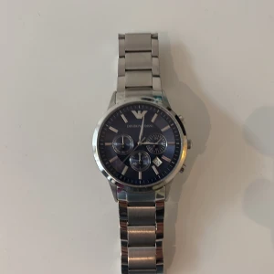 Emporio Armani blå kronografklocka - Snygg kronografklocka från Emporio Armani med marinblå urtavla och silverfärgat armband i metall. Klockan har tre små subdials, datumvisning och klassisk rund form. Perfekt för dig som gillar stilrena accessoarer med lyxig känsla. säljes pga för liten 