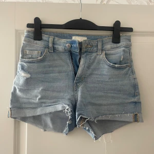 Ljusblå jeansshorts med fransig kant - Säljer ett par ljusblå jeansshorts med hög midja och rå, fransig kant. Shortsen har klassisk femficksdesign, slitna detaljer och uppvikta benslut för en avslappnad look. Perfekta för varma dagar och enkel att matcha med allt.