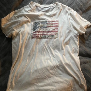 Vit t-shirt Ralph Lauren Denim & Supply - Vit t-shirt från Ralph Lauren Denim & Supply med ett stort amerikanskt flagg-tryck och broderade detaljer på bröstet. Klassisk rund hals och korta ärmar. Tillverkad i 100% bomull för en skön och avslappnad känsla. Om ni har fler frågor är det bara att skriva till mig!!