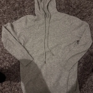 Grå stickad hoodie med huva - Mysig grå stickad hoodie med huva och snörning. Tröjan har långa ärmar och ribbade muddar vid ärmslut och nederkant. Perfekt för kyliga dagar när du vill vara både bekväm och stilren.