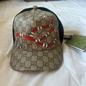 Gucci keps med orm och GG-mönster - Snygg keps från Gucci med klassiskt GG-monogram i beige och brunt, prydd med en röd och vit orm framtill. Baksidan är i svart mesh med justerbart band i grönt och rött. Tillverkad i bomull och polyester, perfekt för dig som vill sticka ut.