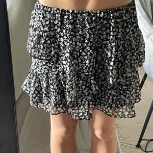 Svart blommig volangkjol - Supersöt svart kortkjol med vita små blommor och volanger. Kjolen har en luftig och flowy känsla, perfekt för varma dagar. Resår i midjan för skön passform och lätt att styla med en enkel topp.