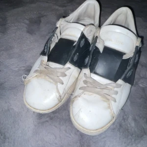 Valentino Open sneakers vit/svart skinn - Säljer ett par Valentino Open sneakers i vitt skinn med bred svart läderdetalj över mitten. Klassisk rund tå, vita snören och platt sula. Snygg och stilren design med diskret Valentino-logga på plösen. Perfekta för dig som gillar exklusiva sneakers med edge.