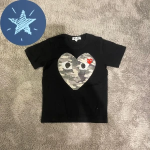 Cdg T-shirt  - Svart t-shirt från Comme des Garçons Play med ett stort hjärta i camouflagemönster och ett mindre rött hjärta med ögon på bröstet. Klassisk rund hals och korta ärmar. Materialet är mjuk bomull, perfekt för en cool och avslappnad stil. Är storlek m men passar s