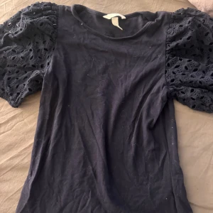 Svart topp med puffärmar och spets - Snygg svart topp med puffiga kortärmar i spets och broderade hålmönster. Kroppen är i mjuk bomull och har rund hals. Perfekt för dig som gillar unika detaljer och vill sticka ut med coola ärmar. Helt ny
