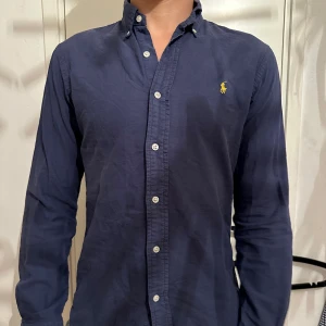 Marinblå skjorta från Polo Ralph Lauren - Säljer en marinblå långärmad skjorta från Ralph Lauren i slim fit. Skjortan har klassisk button-down krage, vita knappar och den ikoniska gula loggan. Storleken är S, modellen är 187