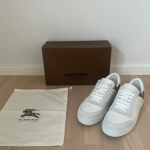 Burberry vita sneakers - Snygga vita sneakers från Burberry med klassisk rutig panel i beige, svart och rött på sidorna. Skorna har ovandel i skinn och mocka, rund tå och vita snören. Perfekta för dig som vill ha en stilren men ändå lyxig look. Kommer med box och dustbag. Dem är helt nya, skriv vid funderingar!