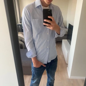 Blå skjorta - Säljer en stilren blåvit randig skjorta från Dressmann i storlek S. Jag tycker att den är lite stor i storleken och jag skulle säga att den sitter som M. Den är i väldigt bra skick. Jag har inte använt den så mycket eftersom att den är lite stor för mig och det är därför jag säljer den.