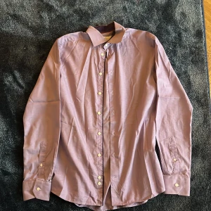 Slim fit rutig skjorta från Eton - Säljer en slim fit skjorta från Eton i storlek 40. Skjortan har ett diskret rutigt mönster i lila och vitt, klassisk krage och vita knappar. Tillverkad i bomull och har långa ärmar med manschett. Perfekt för dig som gillar stilrena detaljer.