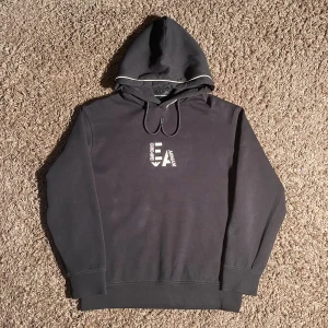 Svart hoodie från Emporio Armani S - Svart hoodie från Emporio Armani med broderad logga i vitt på bröstet. Tröjan har huva med snörning och en diskret vit kant på huvan. Tillverkad i mjuk bomull. Längden är 65 cm och bredden över bröstet är 55 cm.