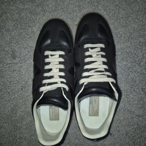 Maison Margiela GATS sneakers svart - SMS FÖR AUTENTISERING. Maison Margiela GATS sneakers i svart skinn och mocka med vita snören och beige sula. Klassisk low-top siluett med rund tå och stilren design. Insidan är vit och skon har märkning på innersulan. Perfekta för dig som gillar minimalistisk och tidlös stil. Har haft dom i några månader och använt dom typ varje dag och dom har endast få tecken på slitningar. 