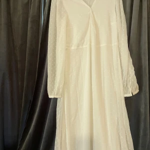 Vit långklänning med prickigt mönster - Lång, luftig vit klänning med diskret prickigt mönster och transparenta långärmar. Klänningen har v-ringning och är tillverkad i ett lätt, flowy material som ger en romantisk vibe. Perfekt för sommarens alla tillfällen.