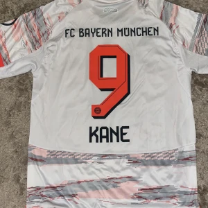Bayern München Kane #9 bortatröja Adidas L - FC Bayern München bortatröja med Kane #9 på ryggen. Adidas Aeroready-material, slim fit. Vit bas med grå och röda grafiska mönster, svarta detaljer och klubbmärke på bröstet. Klassiska Adidas-ränder på axlarna och stor T-Mobile-logga framtill.