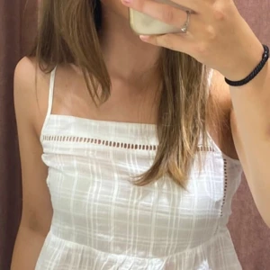 Vit somrig linnetopp med volang - Supersöt vit linnetopp med tunna axelband och volang nertill. Toppen har rutigt vävnad, öppen rygg med knytband. Perfekt för varma dagar och ger en fräsch look till jeans eller kjol💞