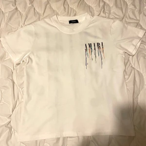  AMIRI t-shirt - Vit t-shirt från AMIRI med tryckt logga och färgstänk-effekt både fram och bak. Klassisk passform med rund hals och korta ärmar. Snygg streetstyle-vibe och tillverkad i mjuk bomull. Perfekt för dig som gillar unika detaljer och vill sticka ut. Använde 4 gånger inget mer! 