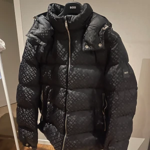 Boss vinter jacka  - Snygg svart pufferjacka från BOSS med diskret rutigt mönster. Jackan har hög krage, dragkedja framtill och två sidofickor med dragkedja. Borttagbar luva och är i knappt använd 