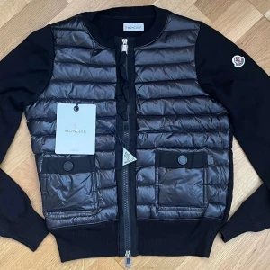 Svart pufferjacka från Moncler - Snygg svart pufferjacka från Moncler med quiltad front i glansigt material och stickade ärmar. Jackan har två stora fickor framtill med knappdetaljer och dragkedja hela vägen. Klassisk Moncler-logga på ena ärmen. Perfekt för dig som vill ha en stilren och exklusiv look. kvitto kommer
