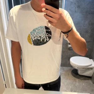 Vit Stüssy t-shirt  - Vit t-shirt från Stüssy i bomull med ett stort grafiskt tryck på bröstet i svart, blått, gult och vitt. Klassisk passform och rund halsringning. Jag är 188cm och bär storlek M. Hör av er vid frågor;)