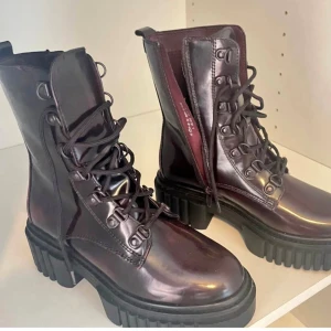 Chunky vinröda boots med snörning - Säljer ett par chunky boots i mörk vinröd färg med grov sula och snörning framtill. Skorna har rund tå och är tillverkade i glansigt skinnliknande material. Perfekta för dig som vill sticka ut med en edgy stil.