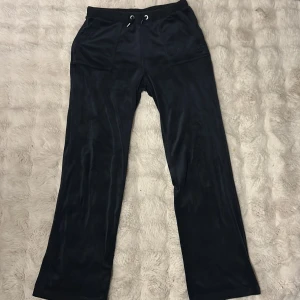 Svarta Juicy Couture mjukisbyxor - Svarta mjukisbyxor från Juicy Couture i mjuk velour med broderad JC-logga och guldfärgade knappar bak. Köpt på Pondus. Byxorna har snörning i midjan, raka ben och praktiska fickor fram och bak. Perfekta för en chill och trendig look. Stl L 