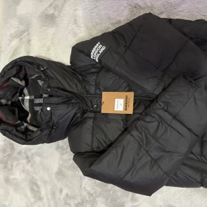 Svart pufferjacka från Burberry M - Svart pufferjacka från Burberry med huva och klassiskt rutigt foder. Jackan har dragkedja framtill, quiltad design och logga på axeln. Materialet är bomull med detaljer i polyester och polyamid. Perfekt för kalla dagar och riktigt snygg streetstil.
