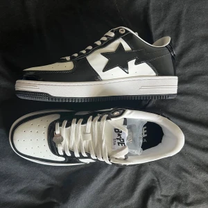BAPE STA svart-vita sneakers - Säljer ett par BAPE STA sneakers i svart och vitt med klassisk stjärndesign på sidan. Skorna har snörning, perforerad tå och en platt sula. Tillverkade i blankt syntetmaterial och läderlook för en clean och stilren vibe. Perfekta för dig som gillar streetwear.