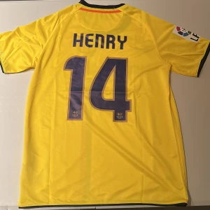 FC Barcelona Henry 14 gul matchtröja - Gul FC Barcelona fotbollströja från Nike med tryck 'Henry 14' på ryggen. Priset är lägre pga att det röd/blåa strecket är placerat mer till vänster än det borde vara, samt klubbmärket. Tröjan har klubbmärke, La Liga-patch på ärmen, svart och röd rand på framsidan samt Unicef-logga. Tillverkad i lätt och ventilerande polyester, perfekt för dig som älskar fotboll och Barca. Replika, endast handtvätt gäller!