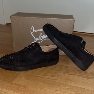 Christian Louboutin svarta sneakers med nitar - Svarta sneakers från Christian Louboutin i mocka med coola svarta nitar på tån. Skorna har snörning och platt sula, samt diskreta detaljer i svart. Perfekta för dig som vill sticka ut med en edgy och lyxig stil.
