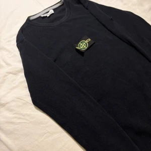 Mörkblå stickad tröja från Stone Island - Säljer en mörkblå stickad tröja från Stone Island med klassisk rund hals och ikonisk logga på ärmen. Tröjan har lång ärm och ribbade muddar för en snygg passform. Perfekt för dig som gillar stilrena och exklusiva plagg.