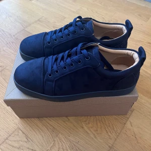 Mörkblå mockasneakers från Louboutin - Snygga mörkblå sneakers från Christian Louboutin i mocka med klassisk röd sula och rund tå. Skorna har blå snörning och beige innersida med guldfärgad logga. Perfekta för dig som vill sticka ut med exklusiva detaljer och lyxig känsla. Använda bara en gång