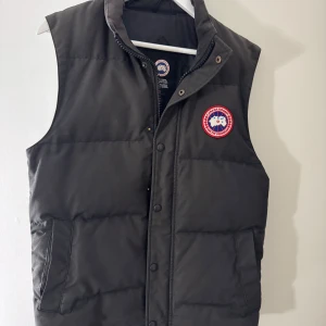 Svart dunväst från Canada Goose - Svart dunväst från Canada Goose med klassisk logga på bröstet. Västen har hög krage, dragkedja och tryckknappar framtill samt två fickor på sidorna. Perfekt för lager-på-lager och riktigt snygg till streetwear-stilen.