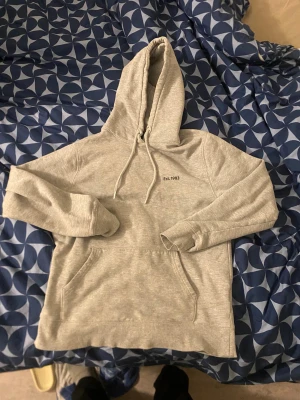 Grå hoodie med tryck och huva - Säljer en grå hoodie med huva och snörning. Framsidan har en liten text 'Est. 1983' och baksidan har ett stort tryck med kors och texten 'Victory Tro Faith Seger'. Tröjan har en klassisk känguruficka och långa ärmar. Perfekt för chill dagar.