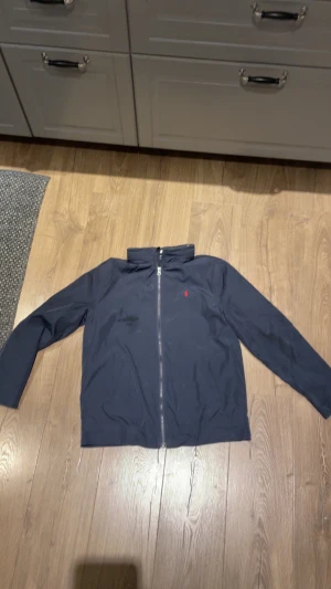 Mörkblå vindjacka från Polo Ralph Lauren - Snygg mörkblå vindjacka från Polo Ralph Lauren med klassisk röd logga på bröstet. Jackan har hel dragkedja framtill, ståkrage och långa ärmar. Perfekt för dig som gillar stilren och enkel design.