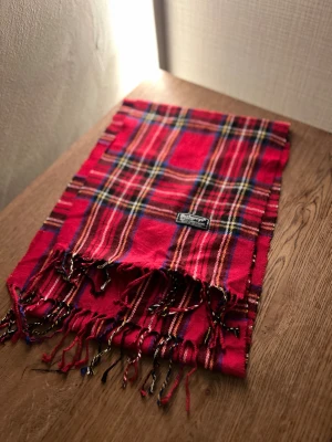 Rutig röd halsduk från Burberry's - Klassisk rutig halsduk från Burberrys i 100% ull. Halsduken har fransar i ändarna och den ikoniska Burberrys-etiketten. Perfekt accessoar för att lyfta din outfit. Anledningen att de stör ”burberrys” är för att de är en äldre modell.