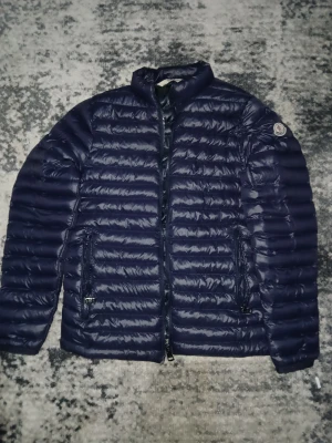 Mörkblå Moncler pufferjacka - Snygg mörkblå pufferjacka från Moncler med klassisk logga på ärmen. Jackan har horisontella quiltade sektioner, dragkedja framtill och två sidofickor med dragkedja. Insidan har coolt serietryck och Moncler-märken. Perfekt för dig som vill ha en stilren och varm jacka.