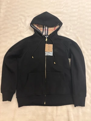 Svart Burberry hoodie med dragkedja - Svart hoodie från Burberry i storlek M med klassisk rutig insida på huvan, guldfärgade detaljer på snörningen och dragkedja. Tröjan har broderad Burberry-logga på bröstet och stora fickor framtill. Perfekt för dig som gillar stilrena och lyxiga streetwear-plagg.
