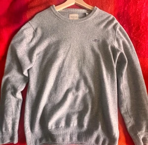 Grå stickad tröja från GANT - Snygg grå stickad tröja från GANT i superfin lammull. Klassisk rund hals och ribbade muddar vid ärmslut och nederkant. Diskret GANT-logo broderad på bröstet. Perfekt för dig som gillar stilrena och bekväma plagg.