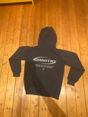Svart hoodie från Chaotic med tryck - Svart hoodie från Chaotic med stor silverfärgad logga och text på ryggen samt mindre tryck på bröstet. Tröjan har huva, ribbade muddar och är långärmad. Perfekt för dig som gillar streetwear och vill sticka ut med coolt tryck.