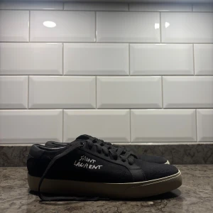 Svarta Saint Laurent sneakers - Snygga svarta sneakers från Saint Laurent med vit logga på sidan. Skorna har snörning, rund tå och en beige sula. Ovandelen är i canvas med detaljer i skinn. Perfekta för dig som gillar exklusiva skor. Pris går att förhandlas vid snabb affär👍