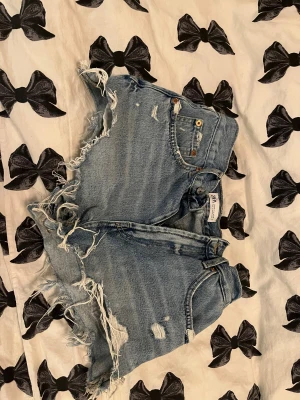 Blå jeansshorts med fransar från ZARA - Säljer ett par blå jeansshorts från ZARA med råa, fransiga kanter och slitna detaljer. Shortsen har fem fickor, knapp och dragkedja framtill samt hög midja. Perfekt för dig som gillar en edgy och avslappnad stil.