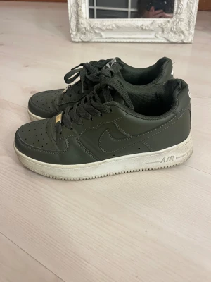 Nike Air Force 1 mörkgröna sneakers - Snygga Nike Air Force 1 sneakers i mörkgrönt skinn med klassisk vit sula. Modellen har rund tå, snörning och perforerade detaljer på tån för extra ventilation. Ikonisk Air Force-logga på plösen och hälen. Perfekt för dig som gillar streetstyle.