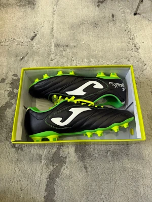 Joma fotbollsskor - Säljer ett par nya Joma fotbollsskor med svart ovandel i syntetmaterial, vita J-loggor på sidorna och limegröna detaljer. Skorna har snörning och gröna sulor med gula dobbar för bra grepp på planen. Insidan är fodrad med grön kant och skorna har en sportig, klassisk look. Storlek 44, köpta för 1499kr säljer just nu för  632kr och är oanvända .