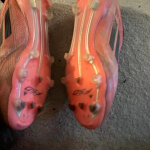 Adidas F50 röda elite fotbollskor  - Säljer ett par Adidas F50 fotbollsskor i röd färg med svarta detaljer och klassiska tre ränder på sidan. Skorna har platt sula med fasta dobbar och är tillverkade i syntetmaterial för lätt vikt och bra grepp på planen. Perfekt för dig som vill ha snabba och snygga skor på matchen. Använda i cirka 2 månader pris kan diskuteras nypris 3100