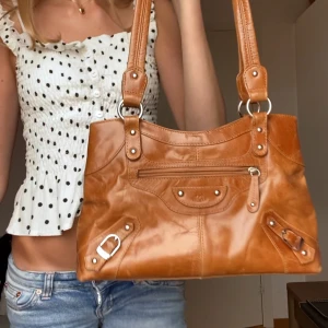 Svin snygg brun skinn axelväska från The Monte (City bag) - Säljer en snygg karamellbrun axelväska från The Monte i äkta skinn. Väskan har silverfärgade metalldetaljer, dragkedja framtill och coola spännen. Perfekt storlek för att få plats med allt du behöver och har två rejäla axelband. Svinsnygg och populär!