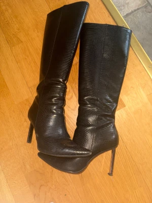 Svarta stövlar Bershka - Säljer ett par svarta stövlar med hög klack och spetsig tå. De har en snygg reptilpräglad yta i skinn och dragkedja på insidan. Perfekta för dig som vill ha en edgy och stilren look med extra höjd.