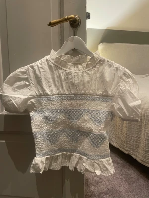 Vit blus med blått mönster och volang - Supersöt vit blus med blåa detaljer från Bik Bok i storlek S med puffiga korta ärmar, volangkant nedtill och smockad midja🩵Den är helt oanvänd varav att lappen är kvar! Hör av dig vid fler frågor!!
