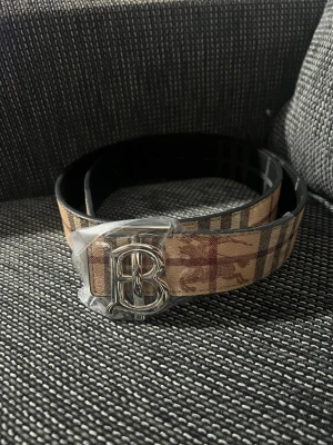Burberry rutig skärp med B-spänne - Säljer ett snyggt Burberry bälte med klassiskt rutig mönster i beige, brun och röd. Bältet har ett stort silverfärgat B-spänne och är tillverkat i skinn. Insidan är svart med präglat Burberry-motiv. Perfekt för dig som vill ha en statement-accessoar.