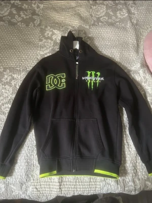 Svart DC x Monster hoodie med huva - Svart hoodie med grön DC och Monster Energy-logga både fram och bak. Tröjan har huva, dragkedja och gröna detaljer vid muddar och nederkant. Perfekt för dig som gillar streetwear och vill sticka ut med coola prints.